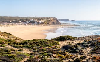 Sweeping panoramic view over Praia de Monte Clérigo beach, capturing the beautiful coastline, blue waters and charming village nestled between the cliffs under a clear blue sky and warm afternoon sun.