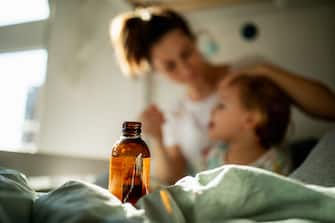 Little girl sitting on her white bed in cozy bright and warm kids bedroom with pastel green background in sunny morning light. Little girl has a cold and her mother is giving her a medicine syrup with a spoon.