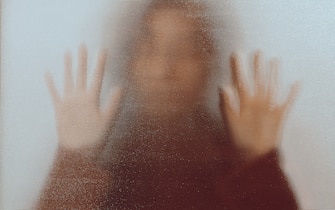 Back lit image of the silhouette of a woman with her hands pressed against a glass window. The silhouette is distorted, and the arms elongated, giving an alien-like quality. The image is sinister and foreboding, with an element of horror. It is as if the 'woman' is trying to escape from behind the glass.