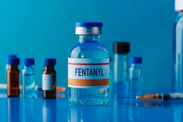 closeup of a simulated vial of fentanyl on a blue table next to a syringe and some other different vials