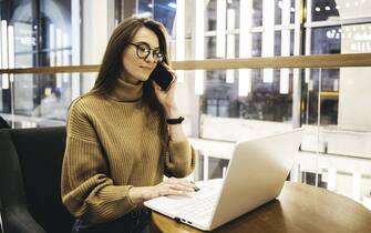 A modern businesswoman in smart AR glasses working from a local cafe with her laptop and wireless digital devices, connected to the internet and making calls. She is wearing a knitted brown wool sweater, sitting at the wooden table. Futuristic dimensional interior, concepts of remote freelance working and women in tech, women career and gender equality