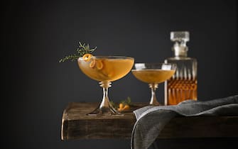 Still life of two Manhattan cocktails garnished with Juniper on a table with whiskey decanter in the background.