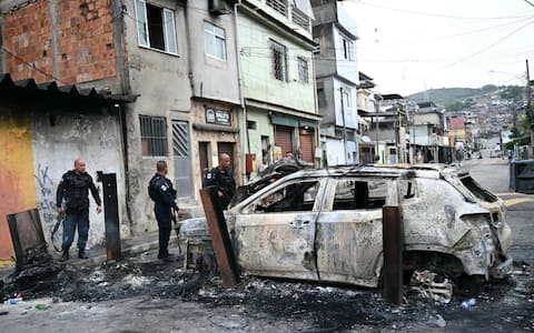 Police officers stand next to a burned car that was part of a barricade set up during the Operacao Contencao (Operation Containment) at the Vila Cruzeiro favela, in the Penha complex, in Rio de Janeiro, Brazil, on October 28, 2025. Hundreds of heavily armed Brazilian police raided slum areas of Rio in a drug raid Tuesday, touching off firefights likened to scenes from a war and leaving at least 64 people dead, Brazilian officials said. (Photo by Mauro PIMENTEL / AFP)