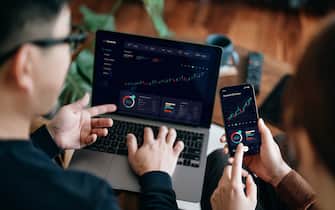 Young Asian couple managing finance and investment online, analyzing stock market trades with mobile app on laptop and smartphone. Making financial plans. Banking and finance, investment, financial trading, mobile banking concept