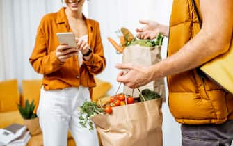 Courier in uniform with thermal bag delivering fresh groceries in paper bags to a client home. Woman with smartphone checking her order