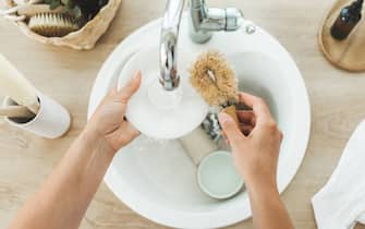 Scrubwoman washes a plate in the kitchen using eco-friendly brushes. Young woman washing dishes with wooden brush with natural bristles at modern white kitchen scandinavian style. Plastic free washing up in kitchen sink. Zero waste and eco friendly home concept.
