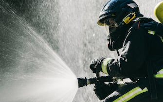 Faceless firemen in safety helmets and protective uniform extinguishing fire with water hose during training on sunny day. Fire extinguishing concept
