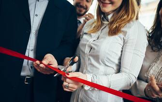 CEO and his assistant cutting a red ribbon, opening ceremony. Few people are in back and they are smiling. They are starting a new business.