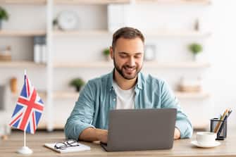 Happy young guy with flag of Great Britain working at desk with laptop in home office. Cheerful male student studying British English online for abroad job, education or emigration