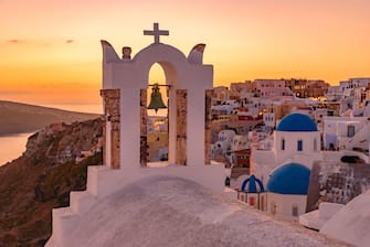 Blue domed churches and bell tower facing Aegean Sea with warm sunset light in Oia, Santorini, Greece