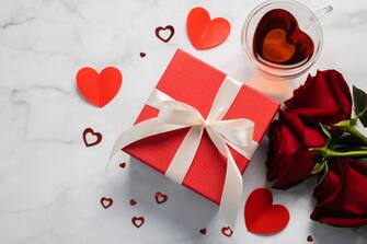 Valentine's Day background. Gift box, red roses, hot tea in cup and aper hearts on background. Flat lay, directly above, copy space.