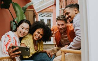 A group of young people sit close together. One woman holds the phone at arms length as she takes a selfie of the group. They all look towards the device and strike a pose.