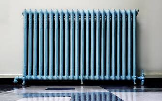 Photograph in Yonkers USA - October 07, 2020. An old hallway cast iron radiator painted blue, with the radiator reflection on the floor tiles.