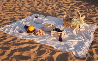 Picnic blanket on the sandy sea shore with picnic basket with flowers, fruits, drinks, bread for an evening picnic. Beach background with picnic blanket and basket, copy space.