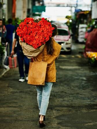 Wide shot rear view of woman carrying large heart shaped bouquet of roses over shoulder after shopping in flower market during vacation