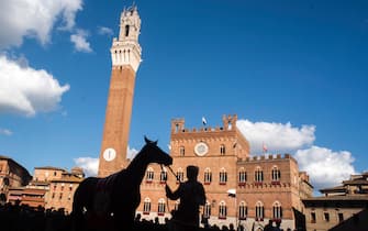 Piazza del Campo – Silhouettes of horse and riders at Piazza del Campo, with Mangia tower in the background. 
Palio, is a horse race that is held twice each year, on 2 July and 16 August, in Siena. Ten horses and riders, bareback and dressed in the appropriate colours, represent ten of the seventeen contrade, or city wards.  
The race itself, in which the jockeys ride bareback, circles the Piazza del Campo, on which a thick layer of earth has been laid. The race is run for three laps of the piazza and usually lasts no more than 90 seconds. It is common for a few of the jockeys to be thrown off their horses while making the treacherous turns in the piazza, and indeed, it is not unusual to see riderless horses finishing the race. Siena (Italy), July 2nd, 2023