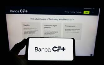 Person holding cellphone with logo of Italian banking company Banca CF+ S.p.A. on screen in front of business webpage. Focus on phone display.