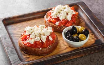 Dakos consists of a slice of cracked barley bread and garnished with crushed or grated ripe tomato and crumbled feta with oregano and olives closeup on the wooden board on the table. Horizontal
