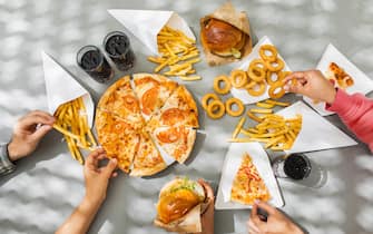 Flat-lay of friends group eating burgers, fries, onion rings, pizza, drinking cola at outdoor party over table background, top view. Fast food dinner from delivery service.Fast food and unhealthy eating concept