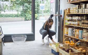 Young white woman opening shop door, she is bending down, placing a doorstop to keep the door open.