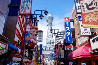 Colorful and showy signs of shops and restaurants against the backdrop of the Tsutenkaku tower in Osaka's Shinsekai entertainment district on a sunny day. Osaka, Japan, late summer 2013.