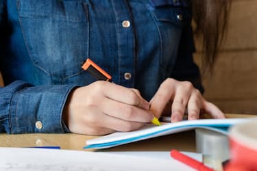 Girl in a blue denim shirt sitting in a cafe makes notes in a notepad with a pen. A sheets with emotions schedule, adhesive tape, felt-tip pens are on the table. Close up