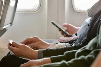 Long shot across laps of older person hands using cell phone.  Framed by seat back and clipped airplane windows.  Hands clearly of senior citizen using cell phone.