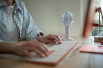 Hands of woman working typing on computer keyboard, small portable compact fan cooling air near on desk. Hot summer weather, lack of air conditioning in office, stuffiness sultriness in workplace.