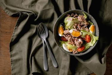 Traditional nicoise salad with canned tuna fish, olives and eggs, served in ceramic bowl with fork and spoon over wooden background with dark linen cloth, Flat lay, copy space. (Photo by: Natasha Breen/REDA&CO/Universal Images Group via Getty Images)