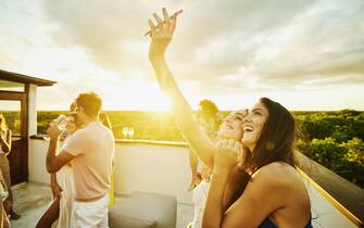 Medium shot of smiling bride taking selfie with friends during rooftop party at sunset after wedding at tropical resort