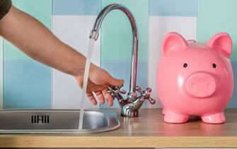 Flowing tap water and a piggy bank standing next to it. Water consumption price concept