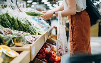 Cropped shot of young Asian woman shopping for fresh organic groceries in supermarket. She is shopping with a cotton mesh eco bag and carries a variety of fruits and vegetables. Zero waste concept