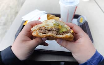 A young man is holding a piece of hamburger in his hands. A bearded guy or man eats fast food. A hungry fat guy is eating an appetizing burger. The concept of junk food, diet, overeating, gluttony, dependence on food. Fast food restaurant, snack.
