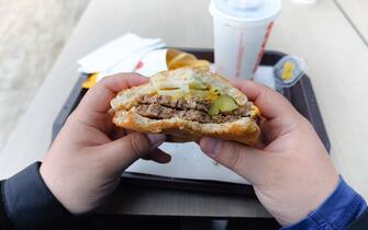 A young man is holding a piece of hamburger in his hands. A bearded guy or man eats fast food. A hungry fat guy is eating an appetizing burger. The concept of junk food, diet, overeating, gluttony, dependence on food. Fast food restaurant, snack.