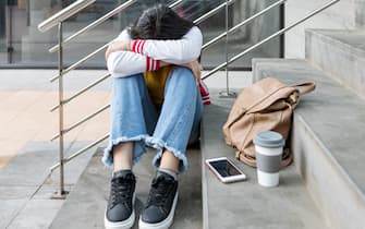 Full body of female student in casual outfit sitting on stairway with smartphone cup and backpack while having problem with bullying