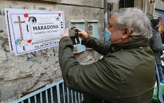 1287832598 NAPLES, ITALY - NOVEMBER 26: An SSC Napoli fan in mourning for Diego Armando Maradona Death take a picture to a death notice poster for Diego Armando Maradona at Quartieri Spagnoli under Diego Armando Maradona mural on November 26, 2020 in Naples, Italy. Diego Armando Maradona died at 60 after a cardiocirculatory arrest while he was at his house in Tigre, Argentina, where he was spending his convalescence after brain surgery three weeks before. (Photo by Francesco Pecoraro/Getty Images)