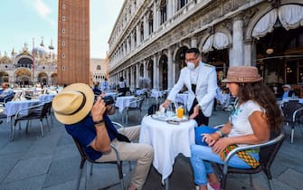 TOPSHOT - A customer (L) takes a photo of a waiter at the terrace of the 18th Century Cafe Florian on June 12, 2020 on St. Mark's Square in Venice, which reopens after being closed for three months during the country's lockdown aimed at curbing the spread of the COVID-19 infection, caused by the novel coronavirus. (Photo by ANDREA PATTARO / AFP) (Photo by ANDREA PATTARO/AFP via Getty Images)