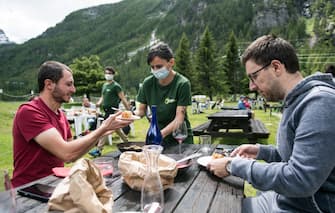 CERESOLE REALE, ITALY - JUNE 14: A waitress wears a protective mask at the mountain refuge to bring food to two men without protective mask sitting at a table near Lake Ceresole Reale inside the Gran Paradiso national park on June 14, 2020 in Ceresole Reale near Turin, Italy. The Gran Paradiso National Park is the oldest national park in Italy, located between the Valle d'Aosta and Piedmont regions, around the Gran Paradiso massif of which the main city is Ceresole Reale. Extending for an area of 71.043,79 hectares on a mainly mountainous terrain it borders on France. After the reopening of commercial activities due to the lockdown for the Coronavirus pandemic, tourism in Italy offers as an alternative to the classic tourist destinations of the beaches also the mountain areas in the Piedmont Region are many tourist areas where you can practice trekking, trekking bicycle, alpine snowboarding or just relaxing for a lunch in a refuge. Italy is returning to a more normal life from easing restrictions after a nationwide lockdown to curb the spread of Covid-19. (Photo by Stefano Guidi/Getty Images)