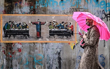 A woman wearing a protective mask walks past a new mural by Italian street artist TvBoy, entitled "Supper for six", inspired by "The last supper" by Italian artist Leonardo da Vinci on October 27, 2020 in Naviglio neighbourhood, southern Milan. (Photo by MIGUEL MEDINA / AFP) / RESTRICTED TO EDITORIAL USE - MANDATORY MENTION OF THE ARTIST UPON PUBLICATION - TO ILLUSTRATE THE EVENT AS SPECIFIED IN THE CAPTION (Photo by MIGUEL MEDINA/AFP via Getty Images)