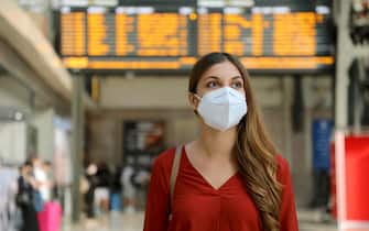 Traveler woman wearing KN95 FFP2 face mask at train station to protect from virus and smog. Young caucasian woman with timetables information of departures arrivals on behind.