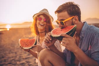 Hipster couple sitting on the beach and eating watermelon. Wearing casual summer clothing, hat and sunglasses. Enjoying in sunset by the sea.