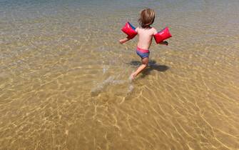 A young boy shot from behind wearing and a blue and red costume and red water wings as a precaution against drowning wades off on his own into a lagoon with a rippled water surface