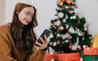 Happy woman reading typing holiday congrats message use smartphone at festive decorated Christmas tree. Smiling beautiful female chatting on mobile