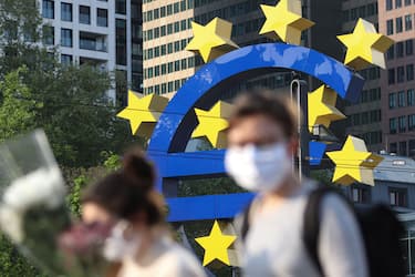 People wearing face masks walk in front of a big Euro sign in Frankfurt am Main, western Germany, as the European Central Bank (ECB) headquarter can be seen in the background on April, 24, 2020 amid the coronavirus COVID-19 pandemic. (Photo by Yann Schreiber / AFP) (Photo by YANN SCHREIBER/AFP via Getty Images)
