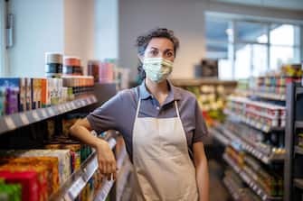 1257380465 Portrait of a woman working in supermarket wearing a face mask looking at camera. Store assistance with face mask and apron leaning to a rack in grocery store.
