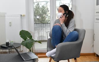 A young woman is sitting in her apartment with a laptop and is working from home because of the corona pandemic, wearing an FFP2 mask and holding a smartphone. MODEL RELEASED! | usage worldwide