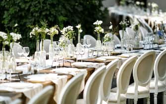 A plates with a light grey napkins are on the table. Nameplate. The table is decorated with white tulle. Also decorated with flower-wooden compositions, glass bottles, simple glasses, white flowers, candles and wooden rack. Fine art. Laid table for a wedding under trees. tables are arranged in an arc. close up of wedding decoration tables outdoor