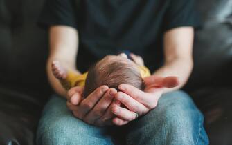 Newborn baby boy with dark brown hair lays cradled in his fathers hands.