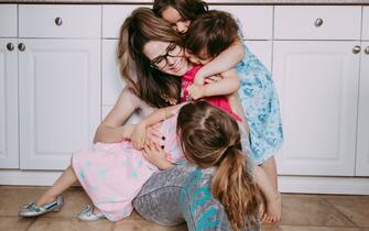 Photo of a young mother sitting on the floor in her kitchen, being surrounded and hugged/loved by her 3 young daughters.