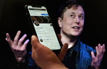 In this photo illustration, a phone screen displays the Twitter account of Elon Musk with a photo of him shown in the background, on April 14, 2022, in Washington, DC. - Tesla chief Elon Musk has launched a hostile takeover bid for Twitter, insisting it was a "best and final offer" and that he was the only person capable of unlocking the full potential of the platform. (Photo by Olivier DOULIERY / AFP)
